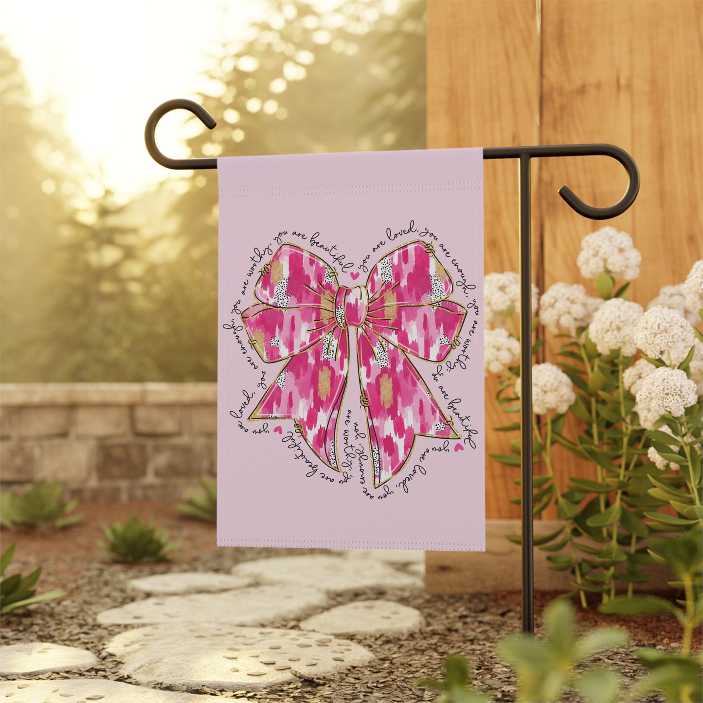 pink bow garden flag with inspirational words you are loved you are worthy you are beautiful in a circle on light pink background, hanging on metal stand in sunny backyard