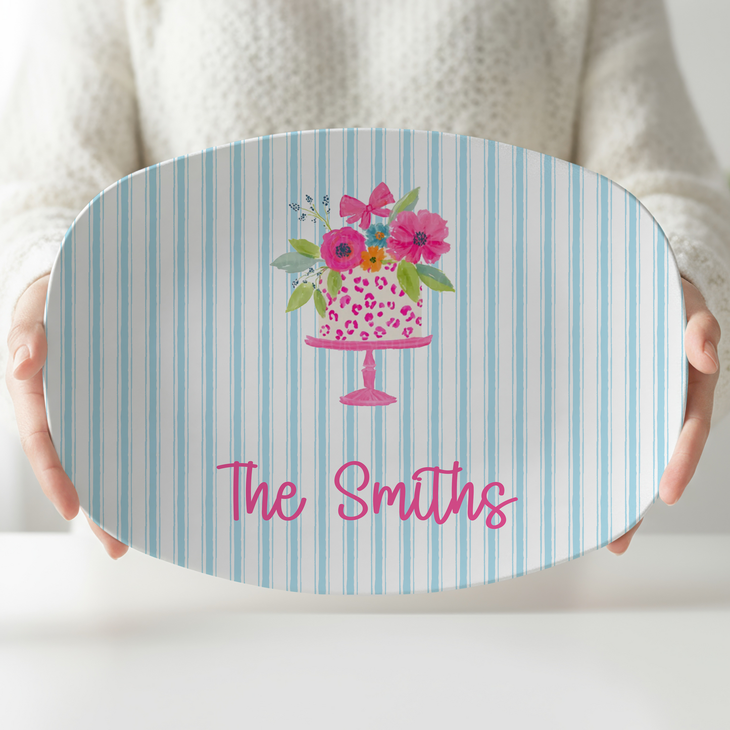 Personalized oval serving platter featuring a watercolor birthday cake with pink flowers, blue ticking stripe background, and a custom family name in bright pink lettering, perfect for birthday celebrations and dessert serving.