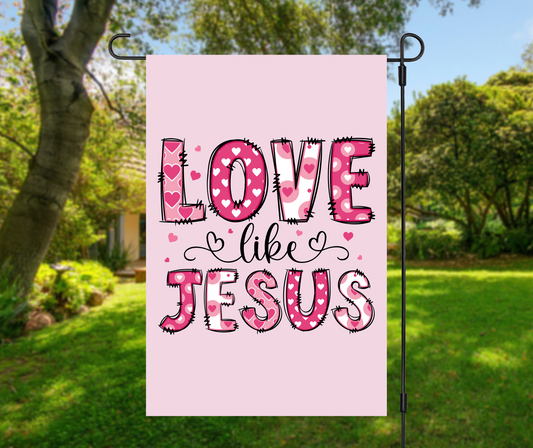 Love Like Jesus garden flag with pink heart letters on light pink background, displayed on metal stand in sunny green yard