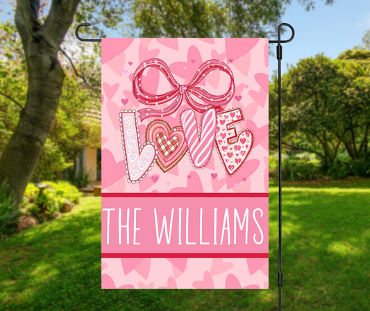 personalized Valentines garden flag with pink LOVE letters, bow and hearts, custom family name THE WILLIAMS, displayed on snowy front porch with flowers